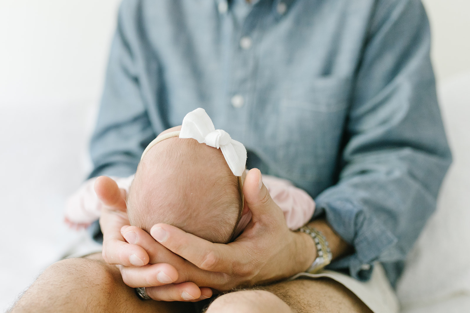 newborn with a white bow being held in the palms of her dad's hands