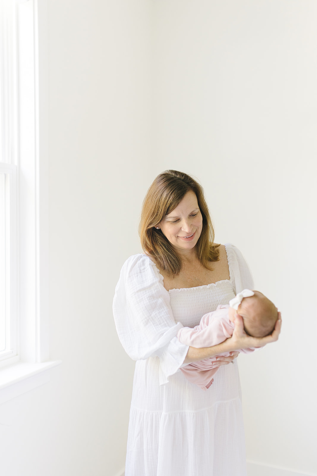 new mom in white dress standing near a window with her newborn Northern Virginia Newborn Photographer