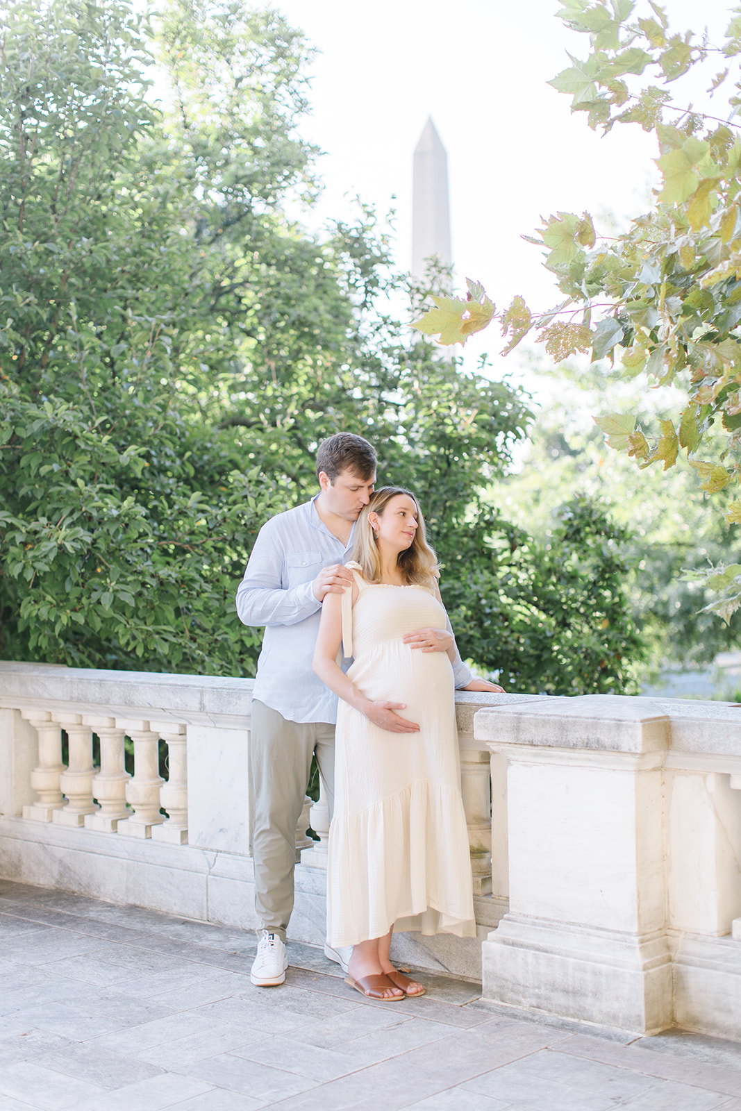 pregnant woman in cream dress holding her bump with her husband kissing her head in front of the washington monument Maternity Photography DC