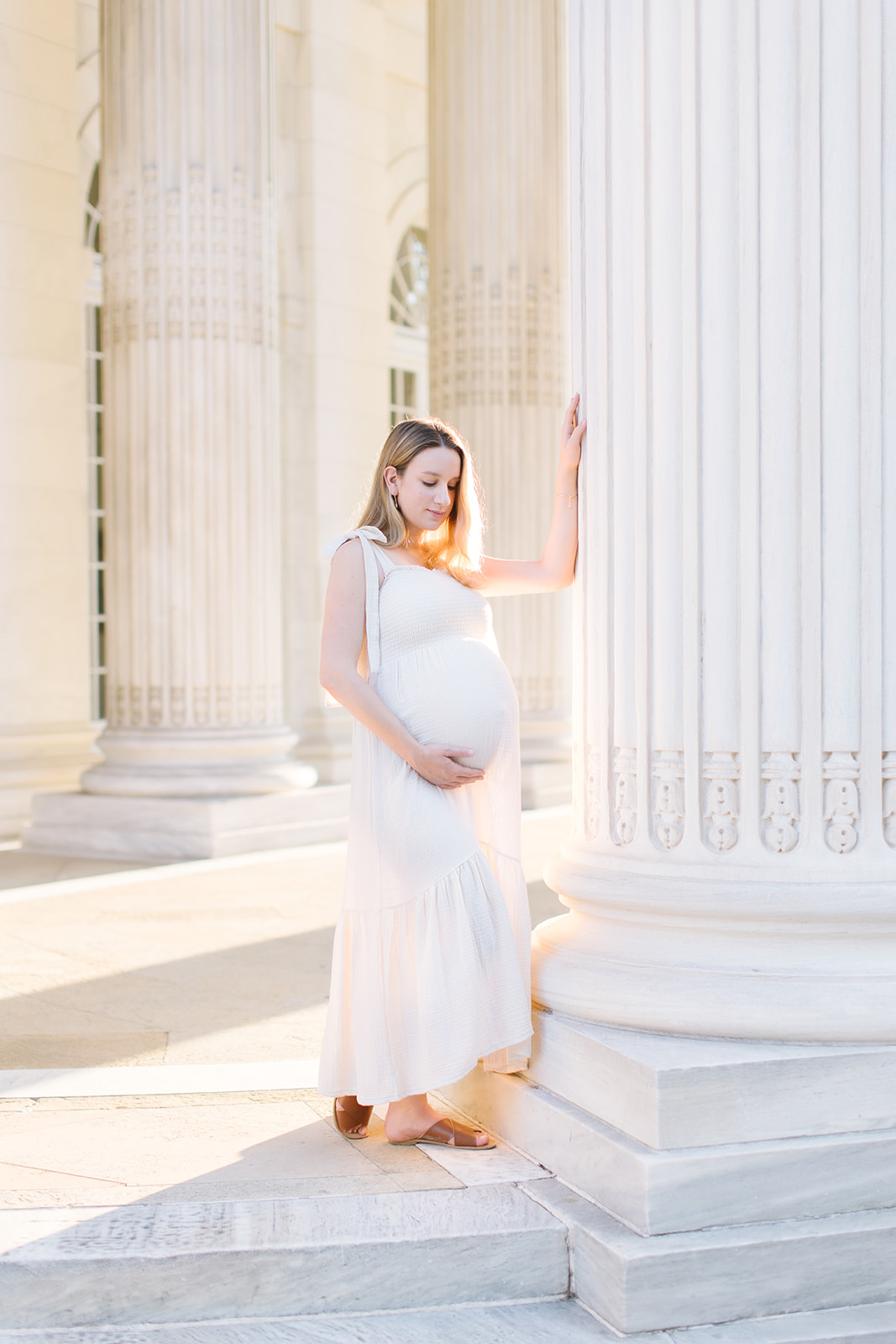 mom to be in a white gown standing near a column in DC Bloom OBGYN