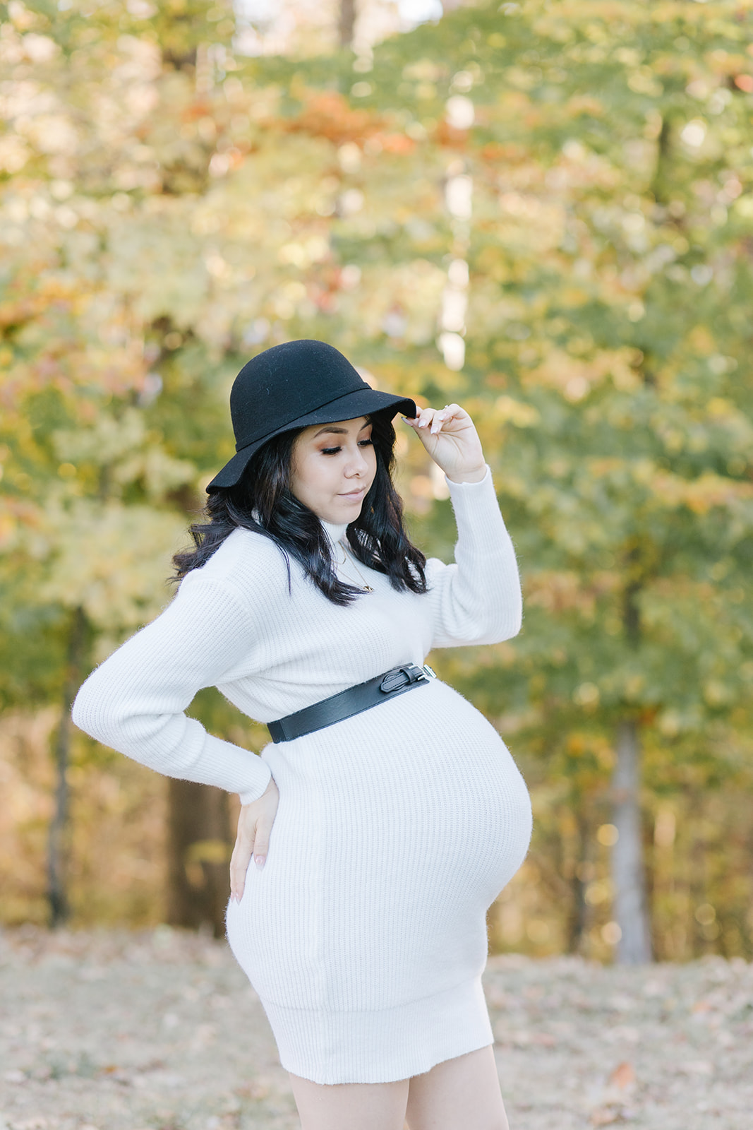 Mom to be in a white dress and black hat with her hand on her back and hat Bloom OBGYN