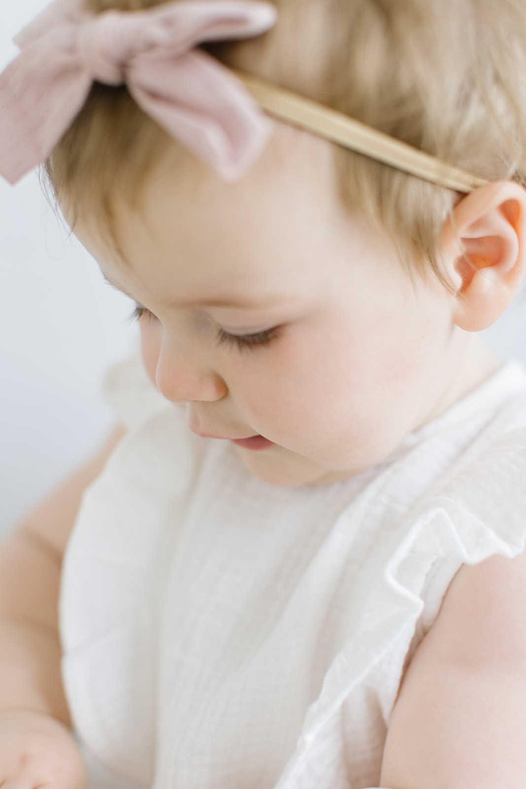 toddler girl looks down at her hands while wearing a bow headband and white dress
