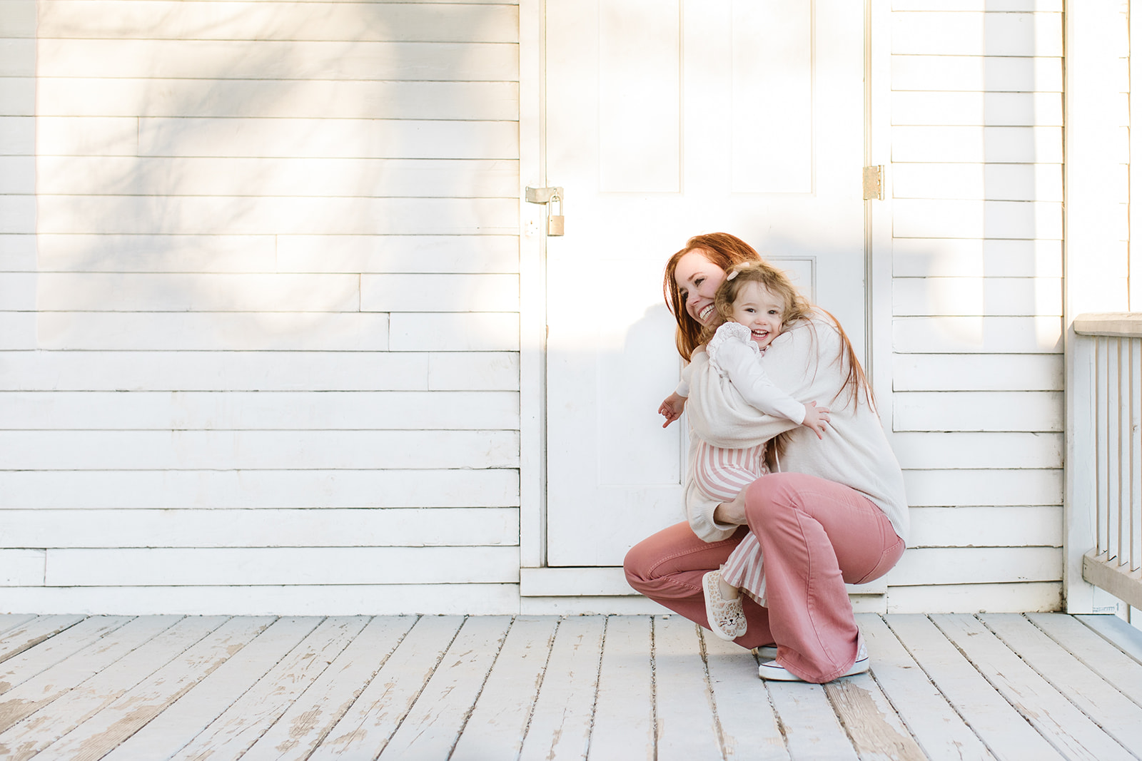 a young girl in a pink striped onesie gets hugged on a white porch by her mother