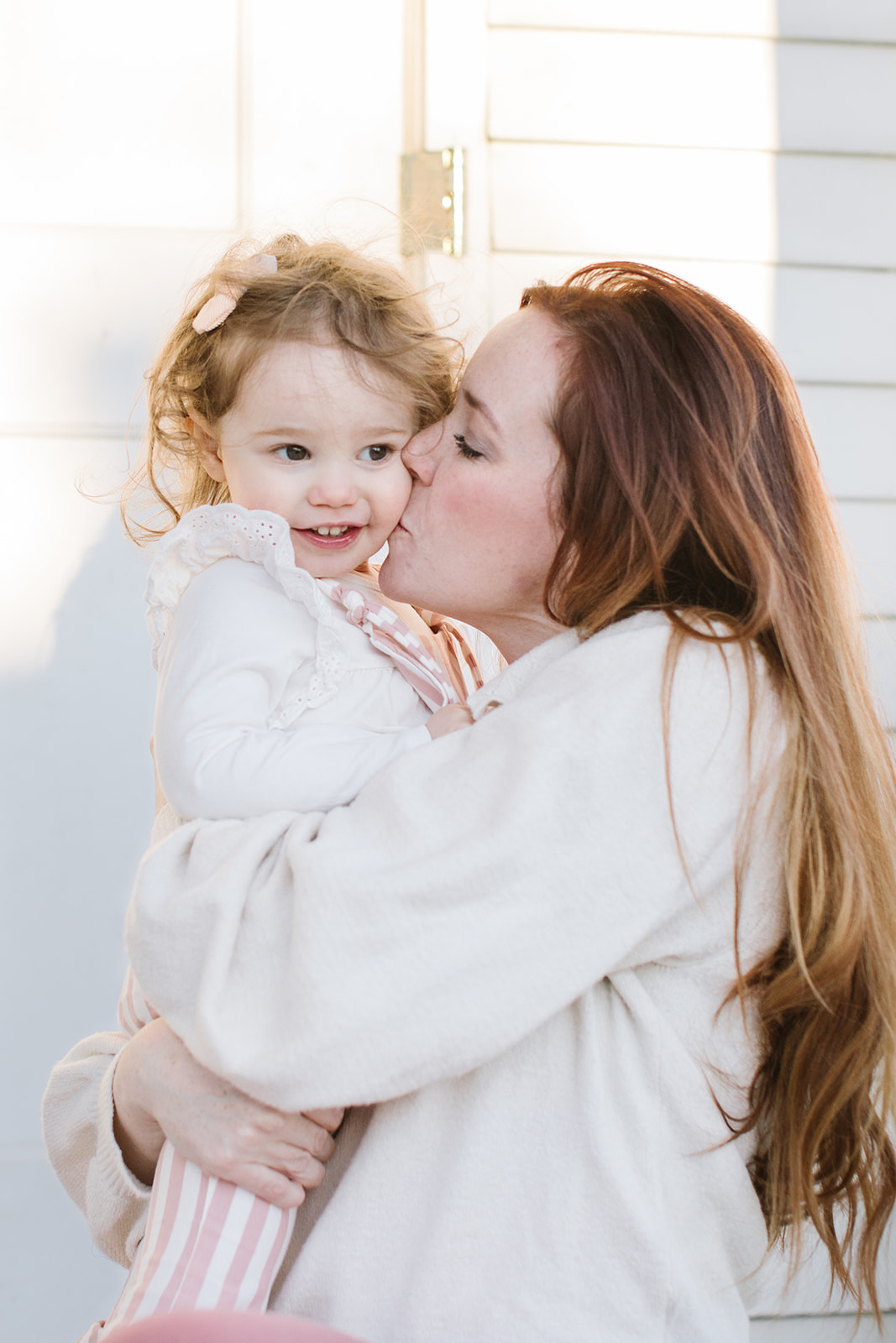 A mother in a white sweater hugs and kisses her young daughter on a front porch nannies arlington va