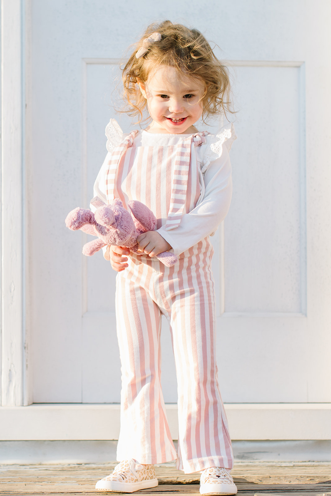 young girl stands in front of a door wearing a pink stripe onesie and holding a pink stuffed animal nannies arlington va