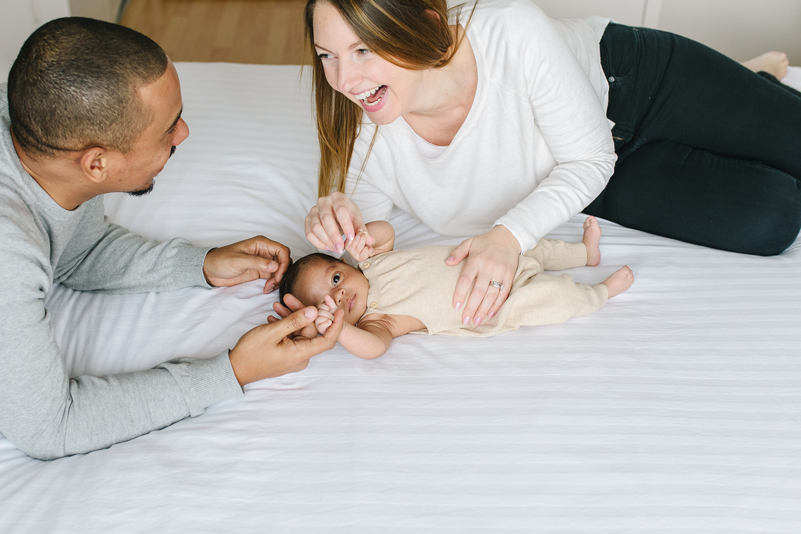newborn baby lays on a white bed in a tan onesie while mom and dad play with it