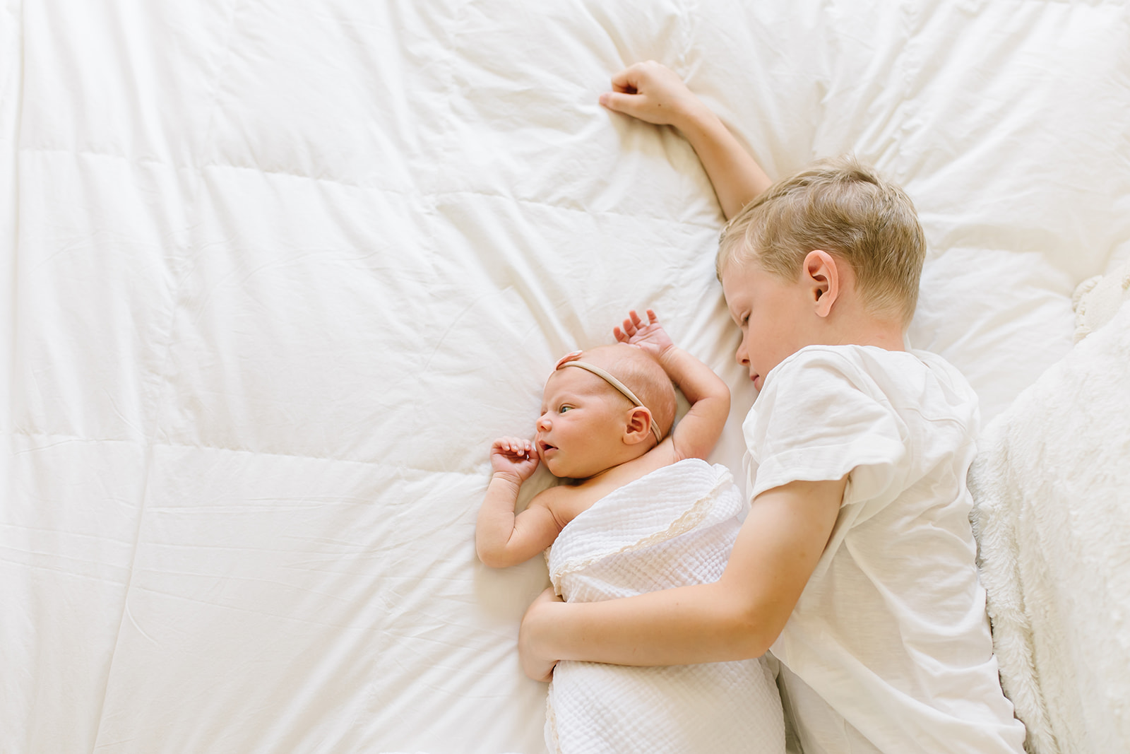 Young boy lays and cuddles with his newborn baby sister on a white bed