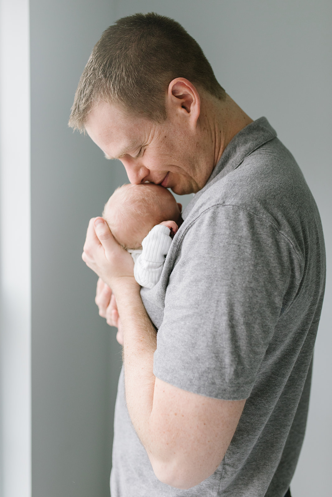 dad stands in front of a window nuzzling his newborn baby in a grey polo shirt